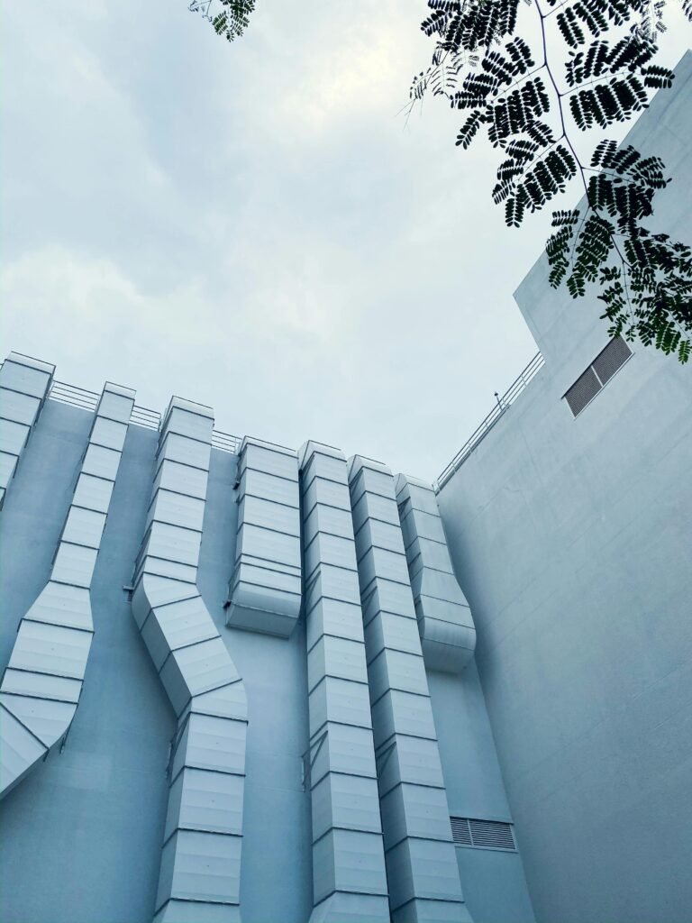 Low-angle view of industrial ductwork on a modern building exterior with sky and tree leaves.